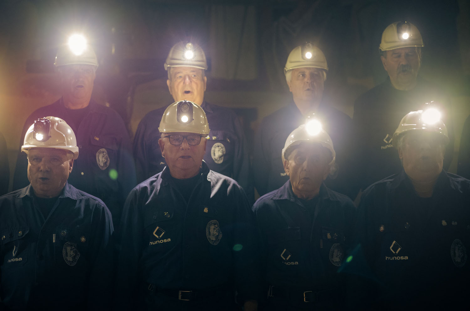 8 men singing in a choir in hard hats with headlamps lit and blue coveralls