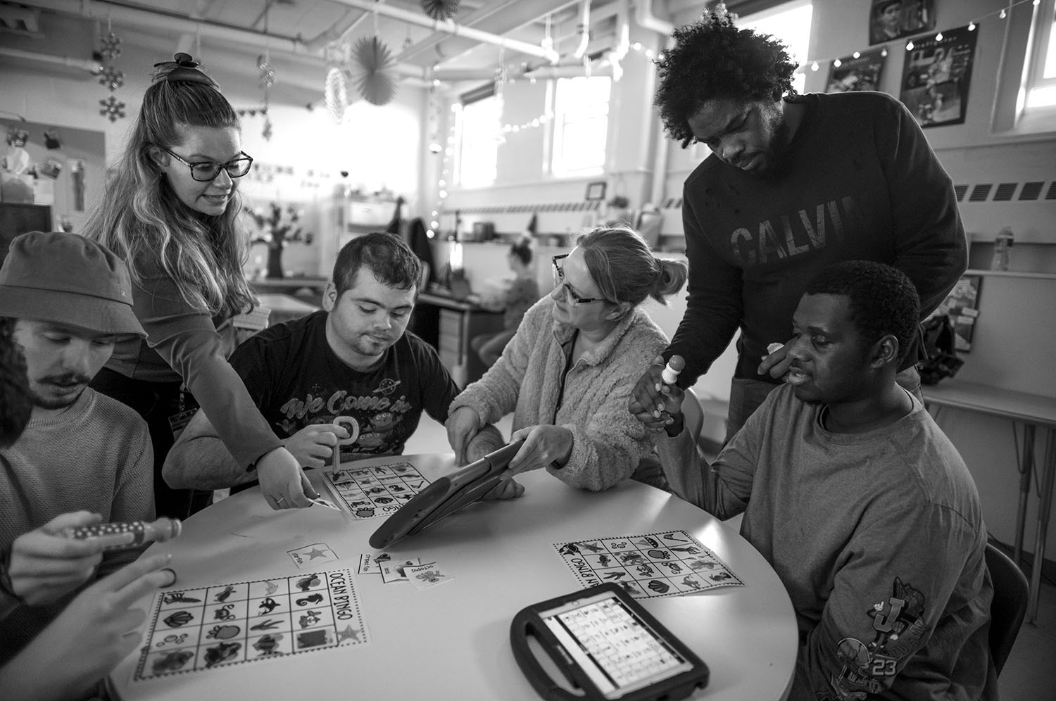 Several people sit around a table and play games. Support staff stand over them.