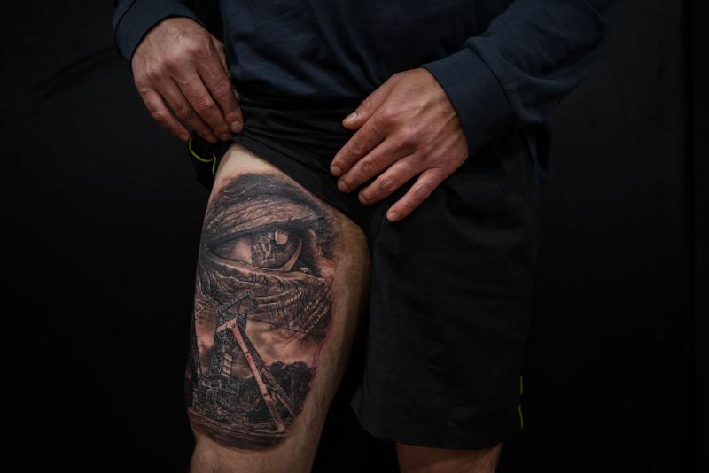 Large thigh tattoo over a man's quadricep. The tatto is an eye above a man digging coal.