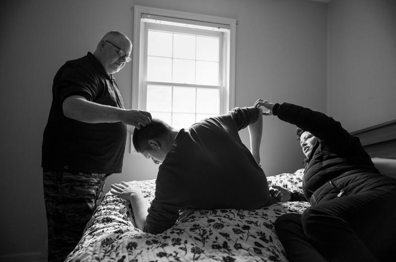 Boy sits on bed; parents' hands on his head and arm.